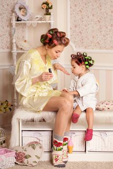 Adorable little girl with her mother in curlers paint their fingernails. Copies mom's behavior. Mom teaches her daughter to take care of herself. Beauty day.