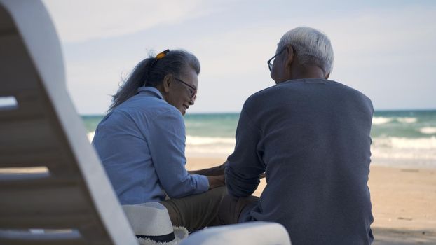 Happy Asian family, senior couple sitting on chairs with backs on beach travel vacation talking together, Romantic elderly enjoy Travel summer vacation, plan life insurance retirement couple concept