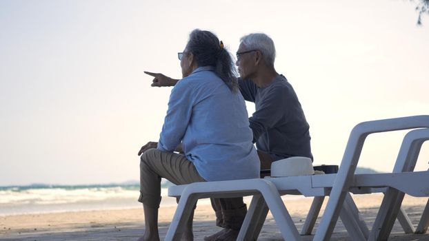 Happy Asian family, senior couple sitting on chairs with backs on beach travel vacation talking together, Romantic elderly enjoy Travel summer vacation, plan life insurance retirement couple concept