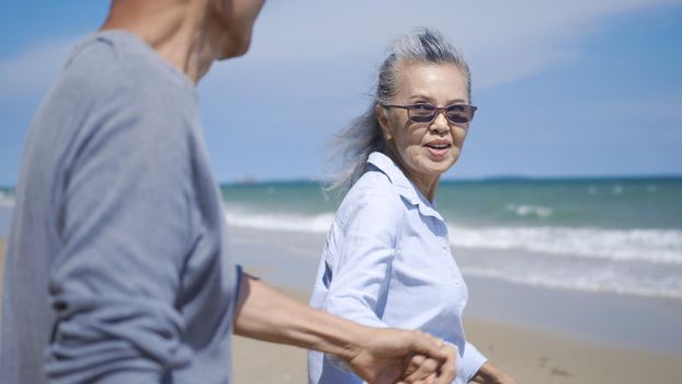 Happy Asian senior man and woman couple holding hands walking to the beach sunny with bright blue sky, Romantic elderly enjoy Travel summer vacation, plan life insurance at retirement couple concept