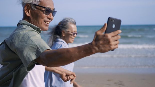 Happy senior couple relaxing outdoors walking on the sea shore and taking selfie together with smartphone on the beach, summer vacation, plan life insurance at retirement couple concept