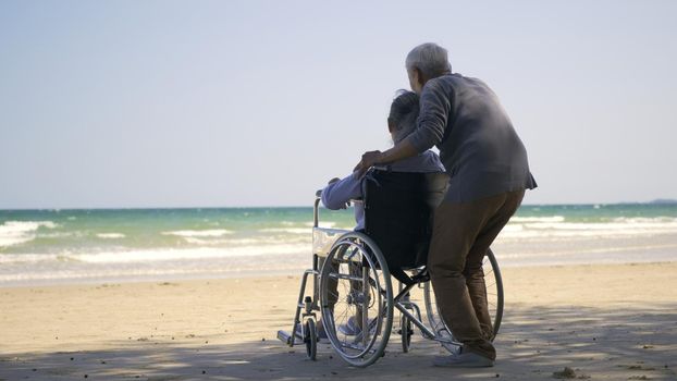 Happy Asian back elderly woman disabled sitting in wheelchair and husband is a wheelchair user on the beach together, summer vacation, Retirement couple concept