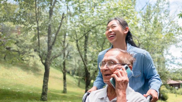 Cheerful disabled senior man talking on smartphone while granddaughter pushing a wheelchair in the park. Family life on vacation.