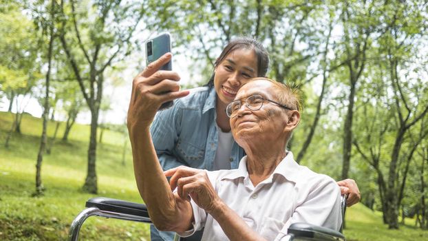 Grandfather in wheelchair and granddaughter talking video call with relatives via smartphone in park. Family life on vacation.