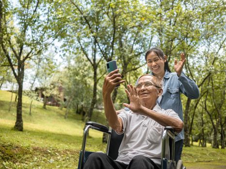 Grandfather in wheelchair and granddaughter talking video call with relatives via smartphone in park. Family life on vacation.