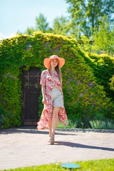 A charming girl in a light summer sundress and a pareo hat is walking in a green park. Enjoys warm sunny summer days.