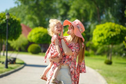 A charming girl in a light summer sundress walks in a green park with her little daughter, holding her in her arms. Enjoys warm sunny summer days.