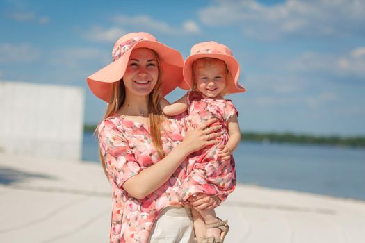 A charming girl in a light summer sundress walks on the sandy beach with her little daughter. Enjoys warm sunny summer days.