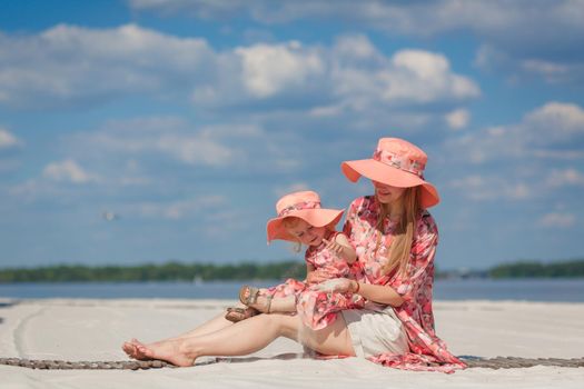 A little girl with her mother in matching beautiful sundresses plays in the sand on the beach. Stylish family look.