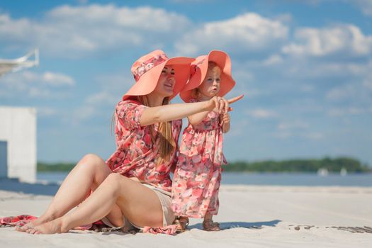 A little girl with her mother in matching beautiful sundresses plays in the sand on the beach. Stylish family look.