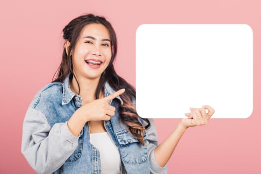 Happy Asian beautiful young woman smiling excited wear denims hold empty speech bubble sign, Portrait female posing finger pointing bubble for your text idea, studio shot isolated on pink background