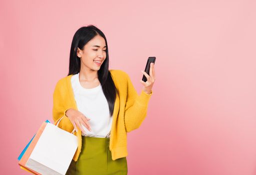 Portrait Asian happy beautiful young woman teen shopper smiling standing excited holding online shopping bags online colorful multicolor and smartphone on hand, studio shot isolated on pink background