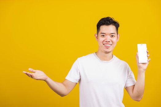 Asian handsome young man smiling positive showing mobile phone blank screen with another hand palm open empty space, excited happy male hold smartphone studio shot isolated on yellow background