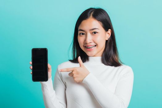 Beautiful Asian woman smile holding a smartphone on hand and pointing finger to the blank screen, female excited cheerful her show mobile phone isolated on a blue background, Technology concept