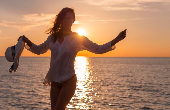 Silhouette of young happy carefree woman posing with hat on the beach at sunset