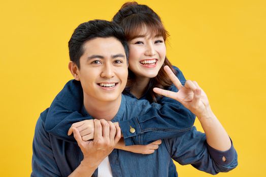 Portrait of happy couple man and woman in basic t-shirts showing peace sign together while doing piggyback ride isolated over yellow background