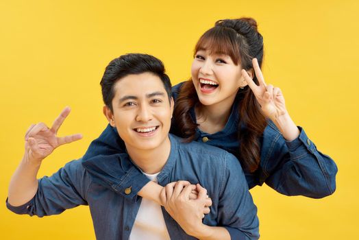 Portrait of happy couple man and woman in basic t-shirts showing peace sign together while doing piggyback ride isolated over yellow background