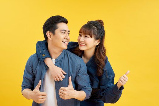 Portrait of smiling couple man and woman showing thumb up together while doing piggyback ride isolated over yellow background