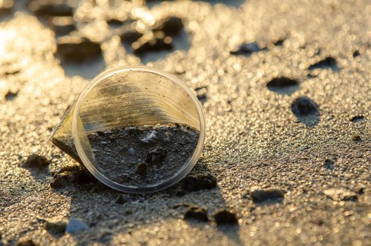 Rubbish plastic cup with sand in morning sunrise at beach. Environmental pollution.