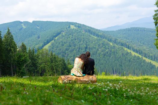 Young couple in love hugs sitting on the log and enjoy betiful of nature mountain lanscape overgrov with green trees.