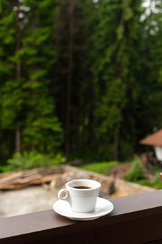 The white mug of dark hot morning coffee on wooden fence with forest background.