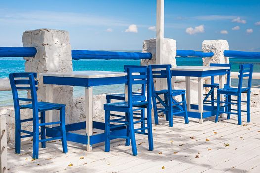 Wooden blue chairs and white tables on seaside terrace with a view on calm waters.