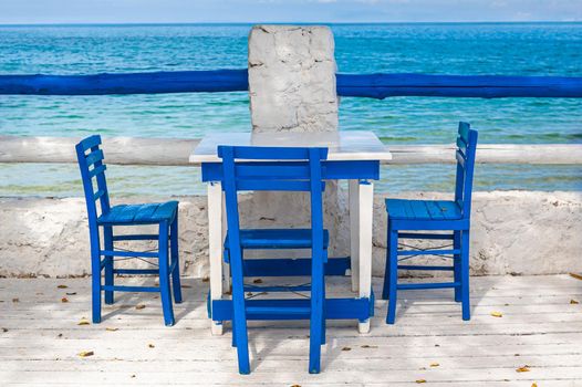 Wooden blue chairs and white tables on seaside terrace with a view on calm waters.