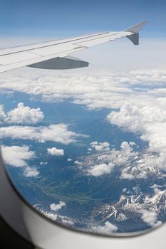 View from airborne airplane window at cloudy sky and Alps mountains with snow covered peaks from high altitude.