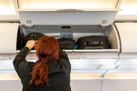 Female passenger reaching out to her luggage in overhead airplane compartment.