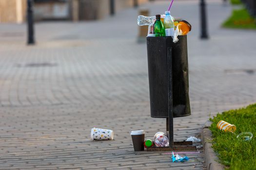 overflowing trash can on the sidewalk pavement at summer daylight.