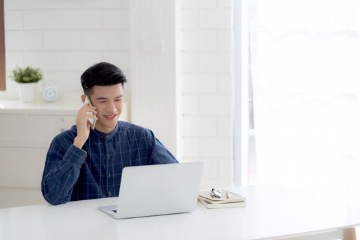 Young asian man talking phone and work from home with laptop computer, freelance using notebook to internet online and speak on smartphone, domestic life, business and communication concept.