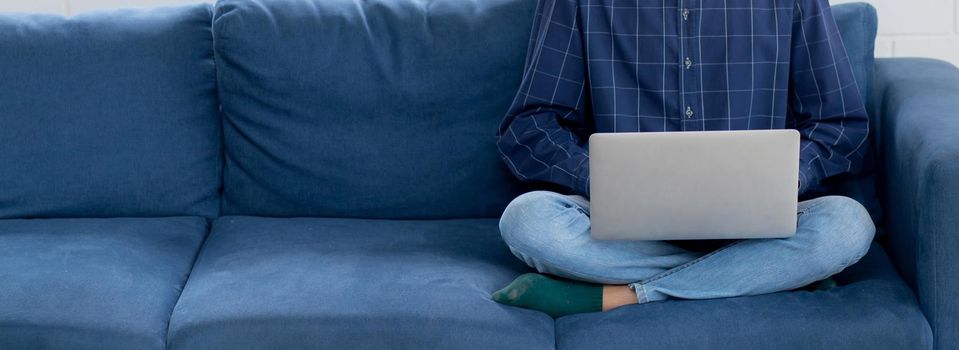 Young asian businessman smile and work from home with laptop computer online to internet on sofa in living room, freelance man using notebook on couch with comfort, new normal, lifestyle concept.