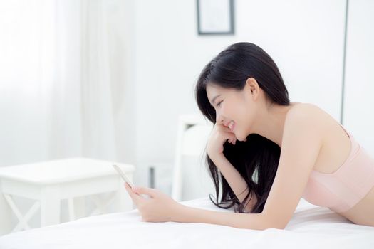 Beautiful young asian woman sexy in underwear chatting on mobile phone in the bedroom, happy girl in lingerie looking social media on smartphone lying on bed, communication and lifestyle concept.