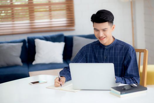 Young asian business man writing on notebook for planning working and using laptop computer on desk at home, notes about finance, male study and learning, business and communication concept.