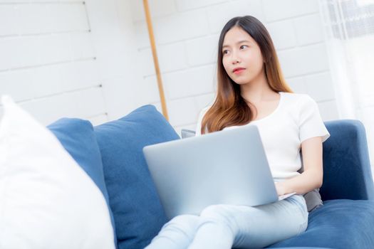 Young asian business woman smile and work from home with laptop computer online to internet on sofa in living room, freelance girl using notebook on couch with comfort, new normal, lifestyle concept.