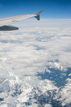 View from airborne airplane window at cloudy sky and Alps mountains with snow covered peaks from high altitude.