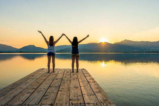 Two girls focused on spiritual meditation looking at setting sun standing on a wooden pier on flat water with light reflections. Suggestive sunset scene of pensive young women in contact with nature