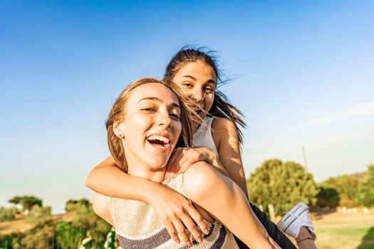 Two girls best friends having fun outdoor in a green nature park looking at camera laughing and joking. Young brunette woman jumping on back of her blonde girlfriend surprising her. Happy students