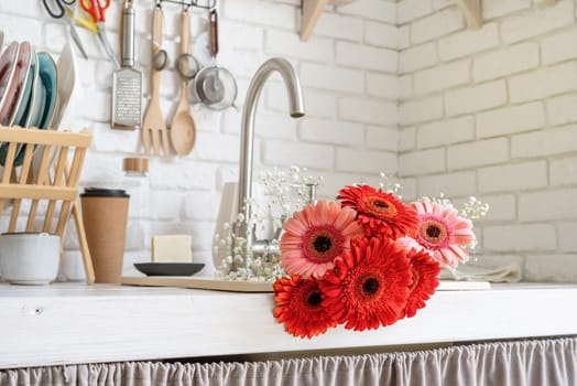 Rustic kitchen interior with white brick wall and white wooden shelves. Red and pink gerbera daisies in a kitchen sink