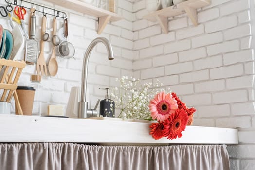 Rustic kitchen interior with white brick wall and white wooden shelves. Red and pink gerbera daisies in a kitchen sink