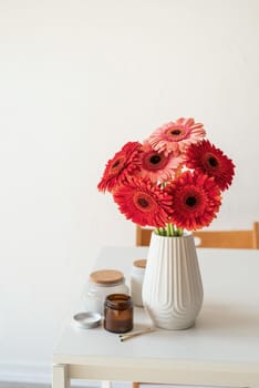 Red and pink gerbera daisies in white vase on table with candles and cup, minimal style