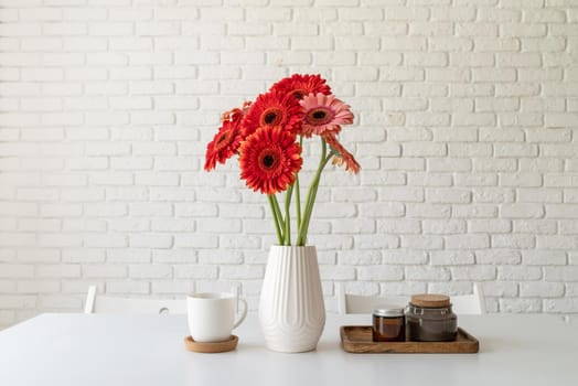 Red and pink gerbera daisies in white vase on kitchen table with candles and cup, minimal style