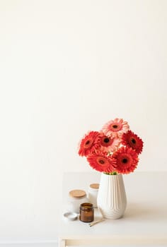 Red and pink gerbera daisies in white vase on kitchen table with candles and cup, minimal style