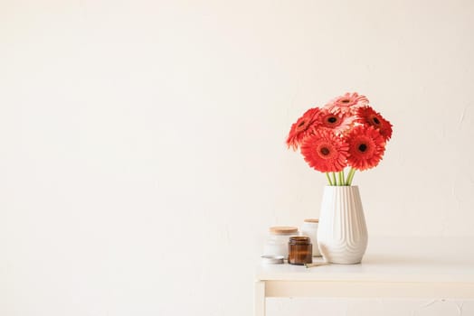 Red and pink gerbera daisies in white vase on kitchen table with candles, minimal style, copy space