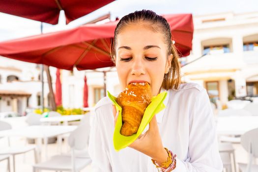 Young hipster millennial cute girl sitting outdoor at bar eating a croissant. Beautiful girl having breakfast in hotel dehors enjoying her vacations. Bad habits of human unhealthy sugar alimentation