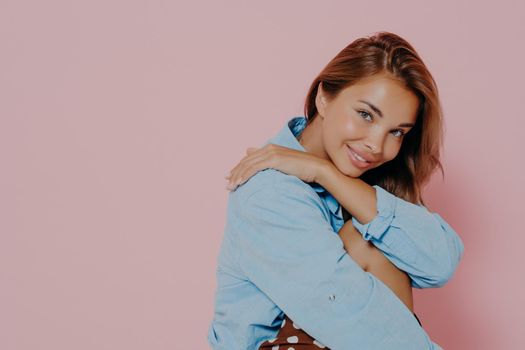 Close up studio portrait of young lovely model female posing seated sideways smiling with slightly tilted head, wearing blue casual shirt, isolated over pastel pink background with blank space