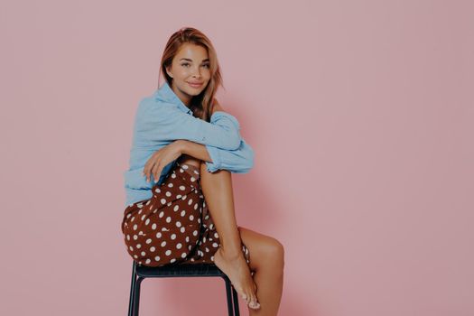Young female model sitting sideways on black modern chair with one leg raised up. Beautiful sensual brunette female in casual shirt and polka dot skirt, isolated over pink studio background