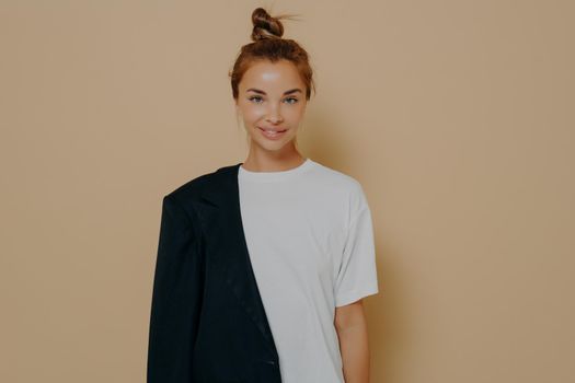 Stylish young female student in white oversized tshirt and black blazer tucked over one shoulder with hair tied up in bun, posing in studio against beige background with blank space smiling on camera