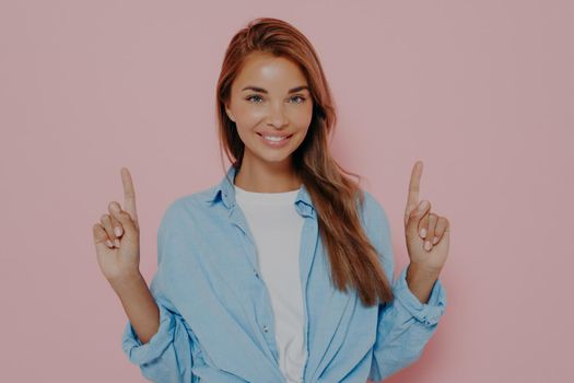 Happy young female points upwards with index fingers with broadly smile, shows awesome item on sale, wears white tshirt with blue shirt, isolated on pink studio wall. Promotion and advertisement