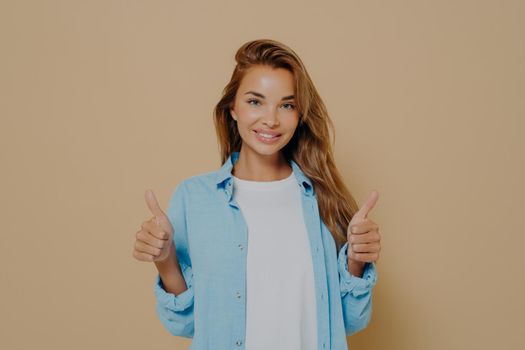Charming young woman showing thumbs up gesture with both hands, demonstrating agreement and approval, smiling at camera isolated over brown background. Body language concept